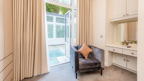 Une pièce avec un fauteuil gris, des rideaux beige, une coiffeuse blanche et une porte vitrée donnant sur l’extérieur.