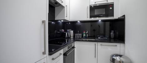 Une cuisine moderne avec des meubles blancs, un plan de travail noir, un four à micro-ondes Bosch, une bouilloire et une poubelle en acier inoxydable.