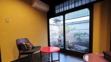 A room with a wooden floor, a red table, a black chair with a cushion, and a view of a garden through a large window.