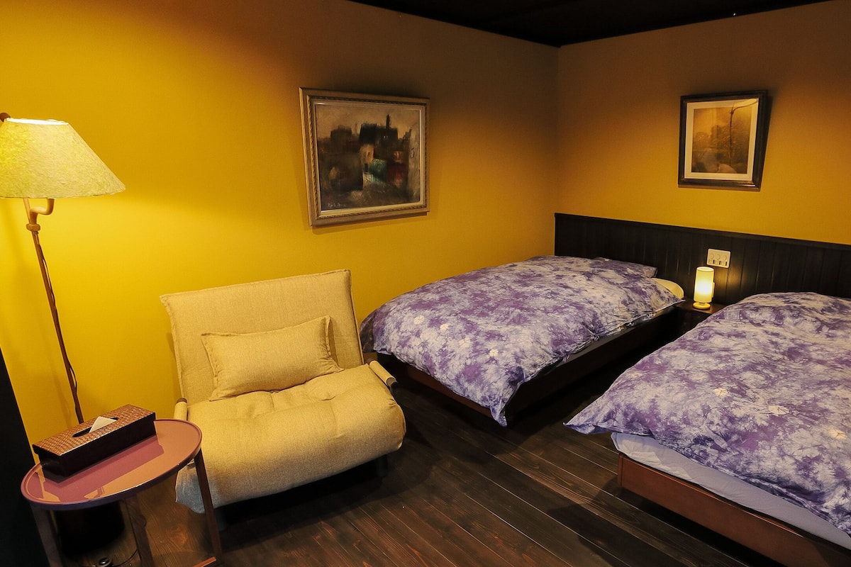 Una habitación de hotel con dos camas, una silla, una lámpara y dos cuadros en la pared.