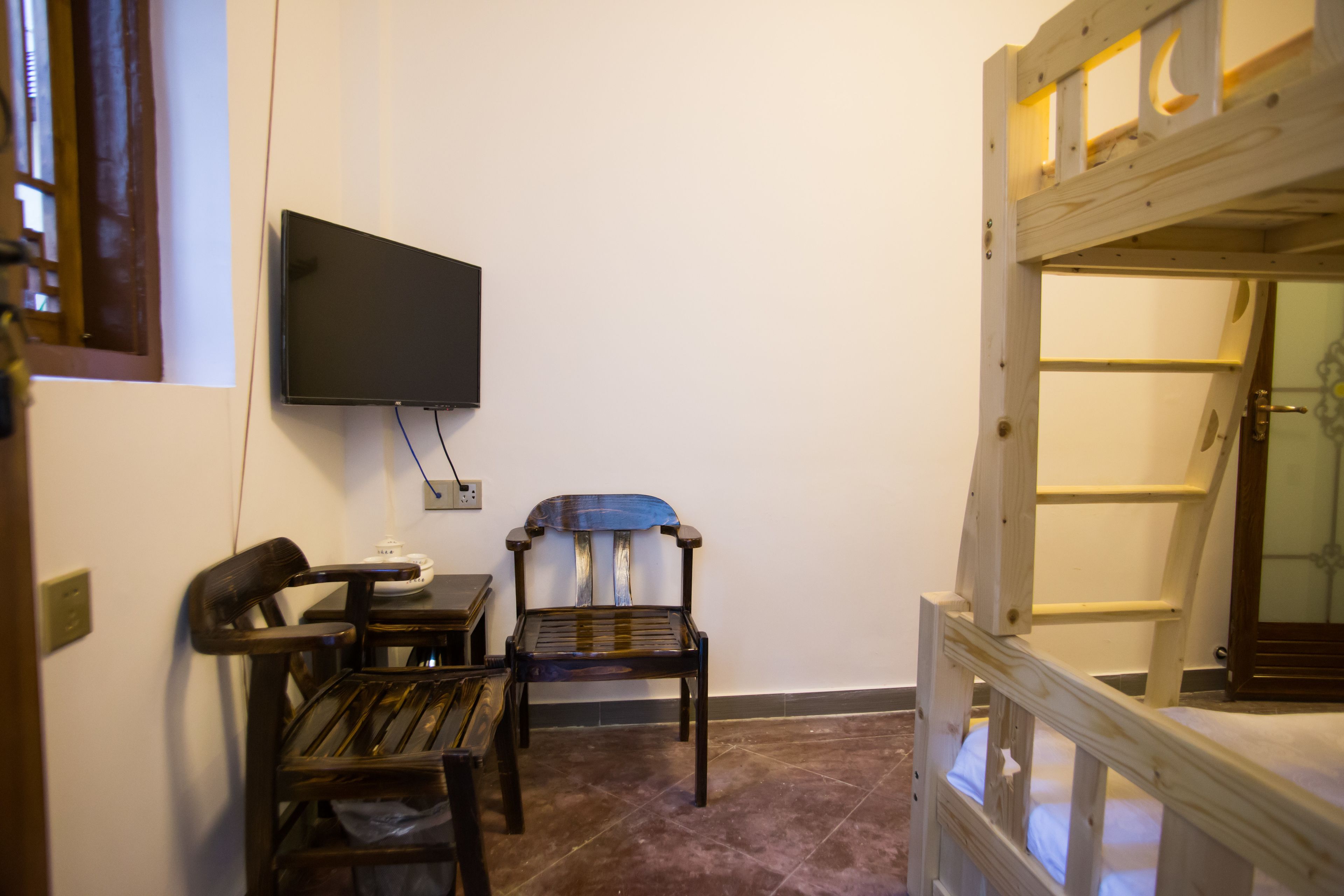 A room with a television mounted on the wall, a wooden chair, a small table, and a wooden bed frame.