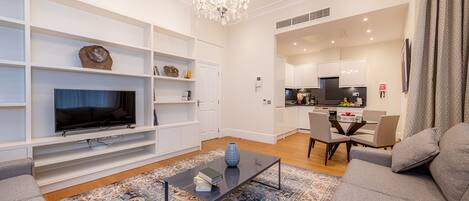 A modern living room with a flat-screen TV, a coffee table, and a sofa. There is a kitchen in the background with a dining table and chairs.