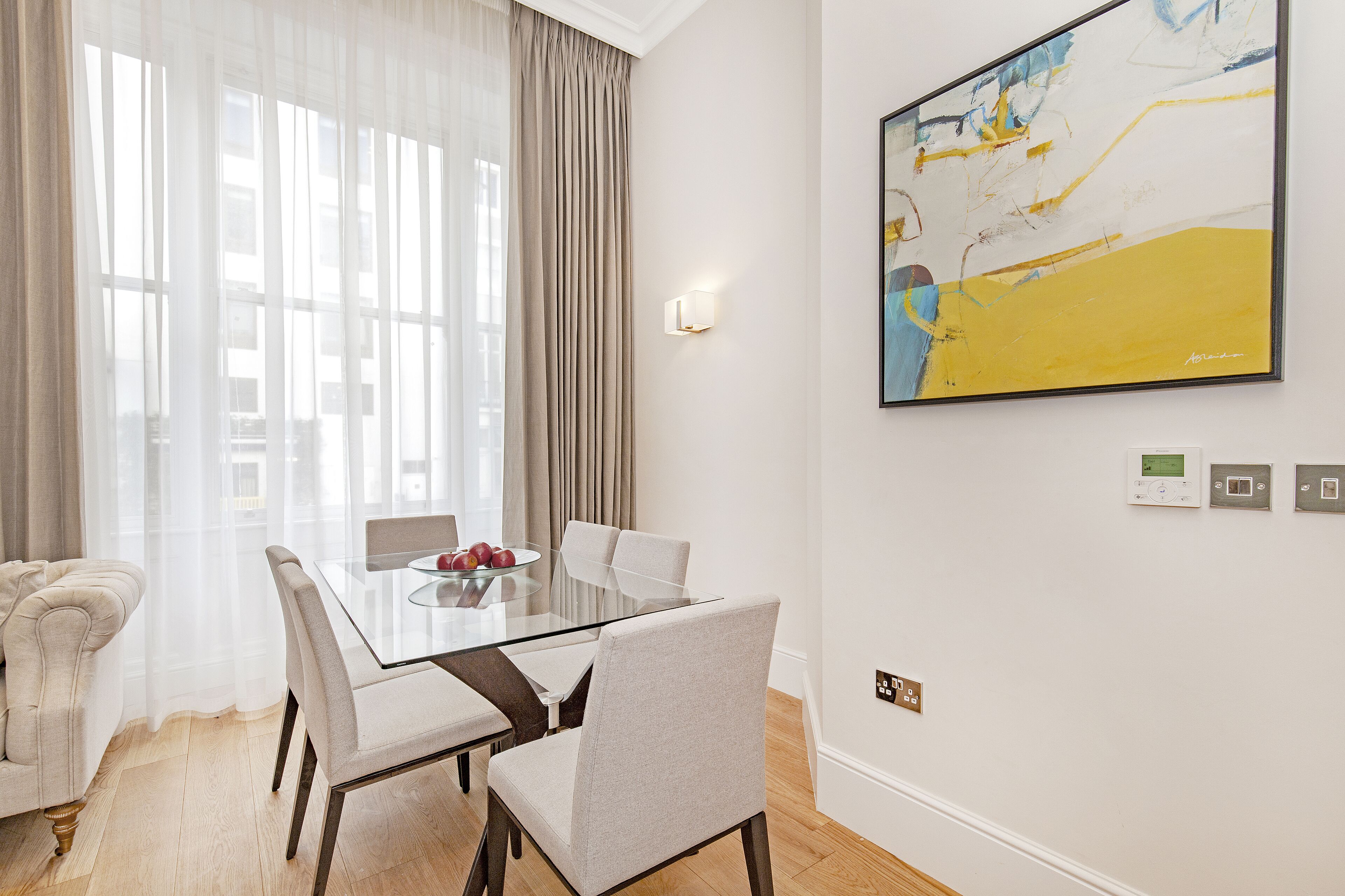 A modern dining room with a glass table, four chairs, a sofa, and a large abstract painting on the wall.