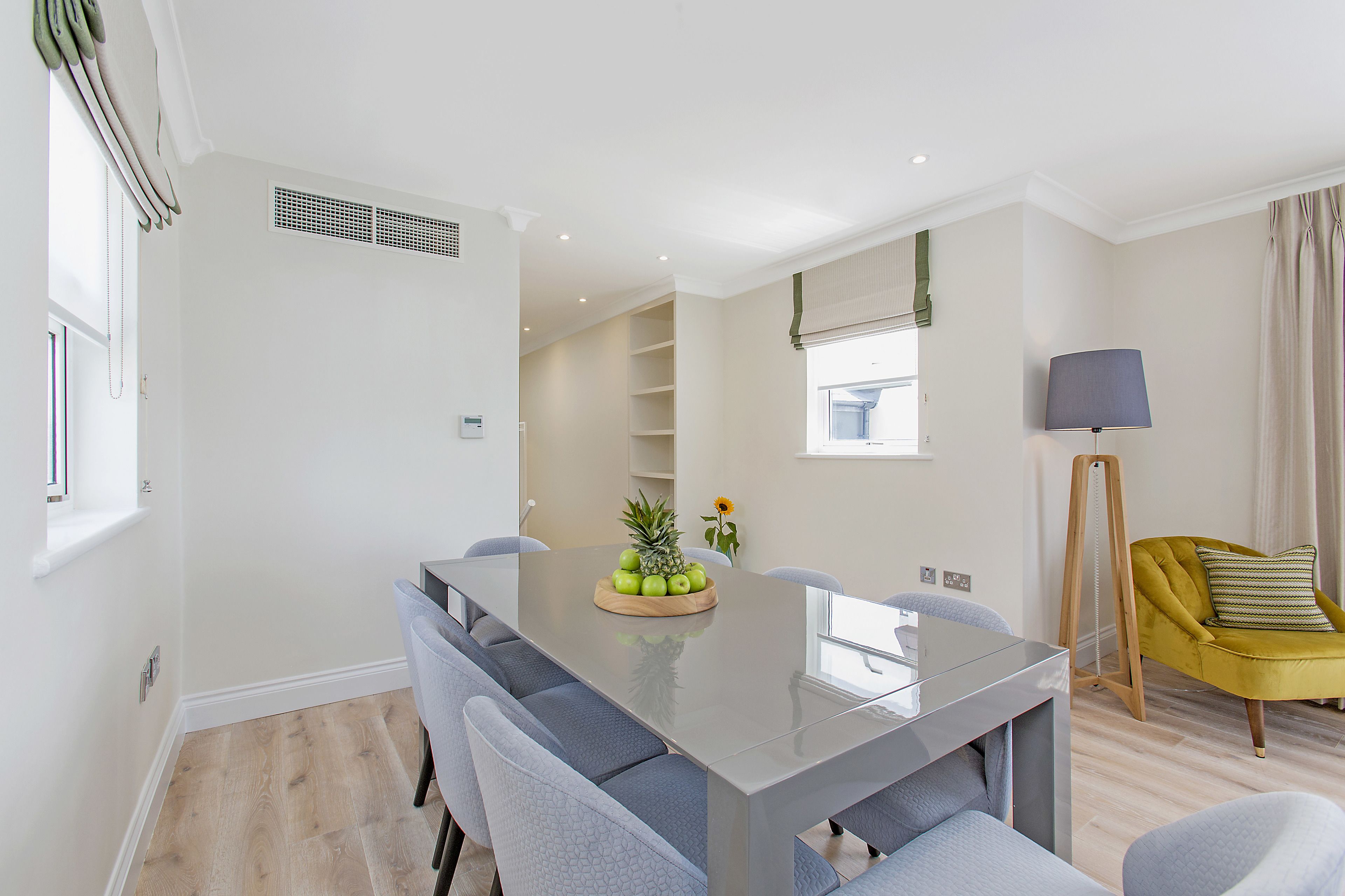 A modern dining area with a glass table, grey upholstered chairs, a wooden side table with fruit, and a yellow armchair with a patterned cushion.