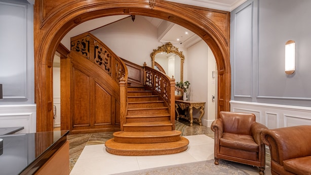 Un magnifique escalier orné de boiseries sculptées, un miroir et un fauteuil en cuir dans un hall d’entrée spacieux.