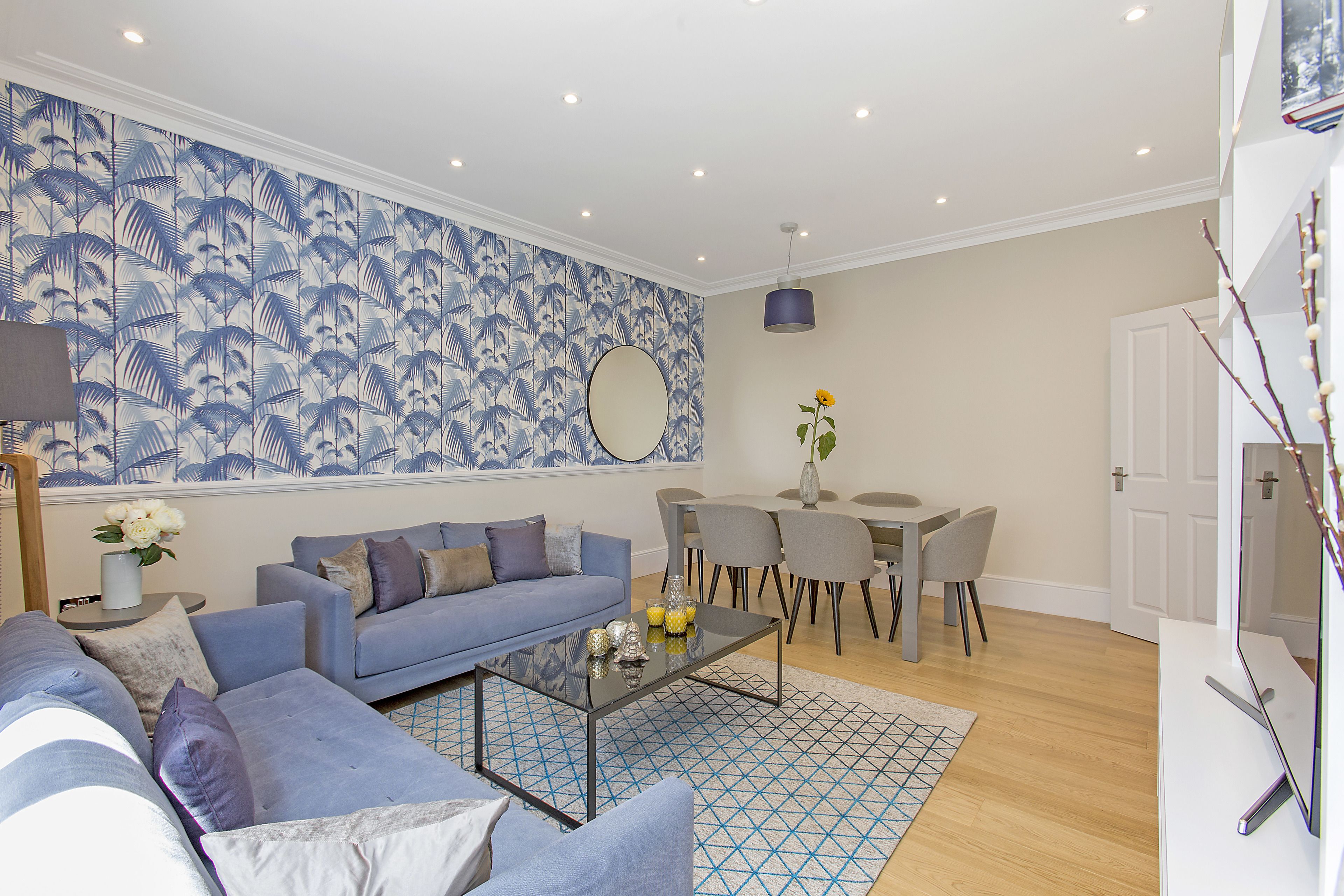 A modern living room with a blue patterned wallpaper, a glass coffee table, a blue sofa, and a dining area with grey chairs and a white table.
