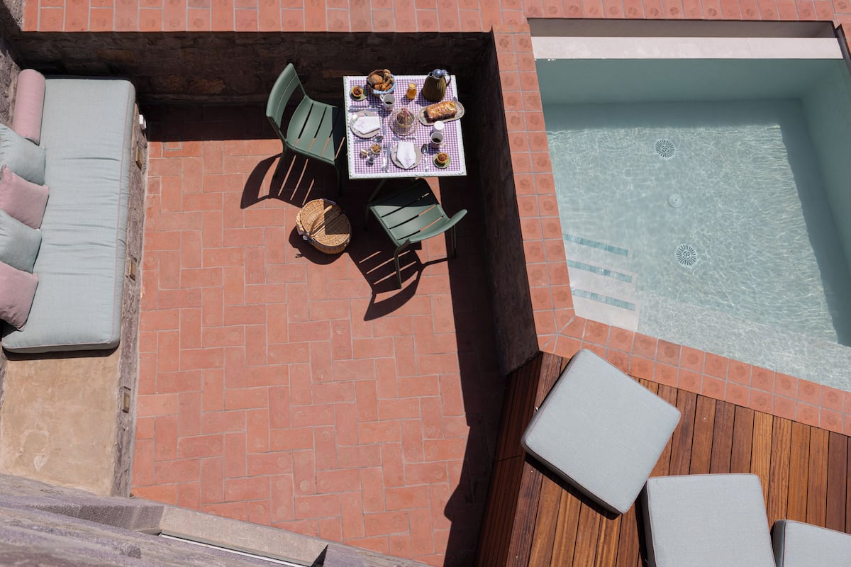 A rooftop pool area with a table set for dining, surrounded by lounge chairs and a sunbed.