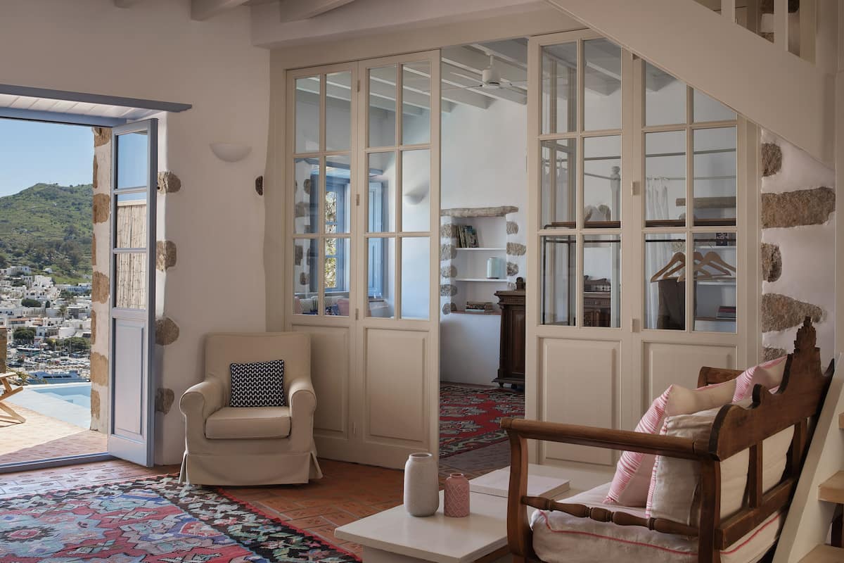 A room with a large sliding glass door leading to a balcony, a wooden chair with a cushion, a small table with a vase, and a bookshelf with various items.
