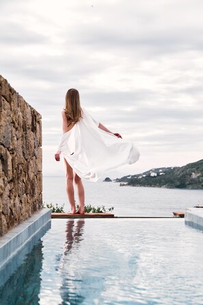 Una persona está de pie al borde de una piscina infinita, con vistas al mar y colinas al fondo.