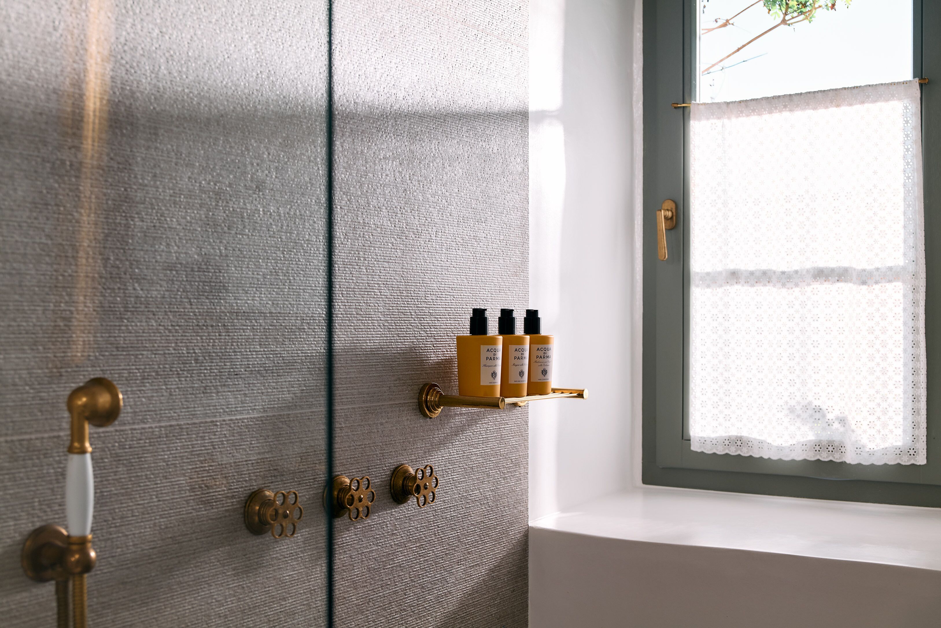 Baño con pared texturizada, picaporte de latón, repisa con botellas y ventana con cortina de encaje.