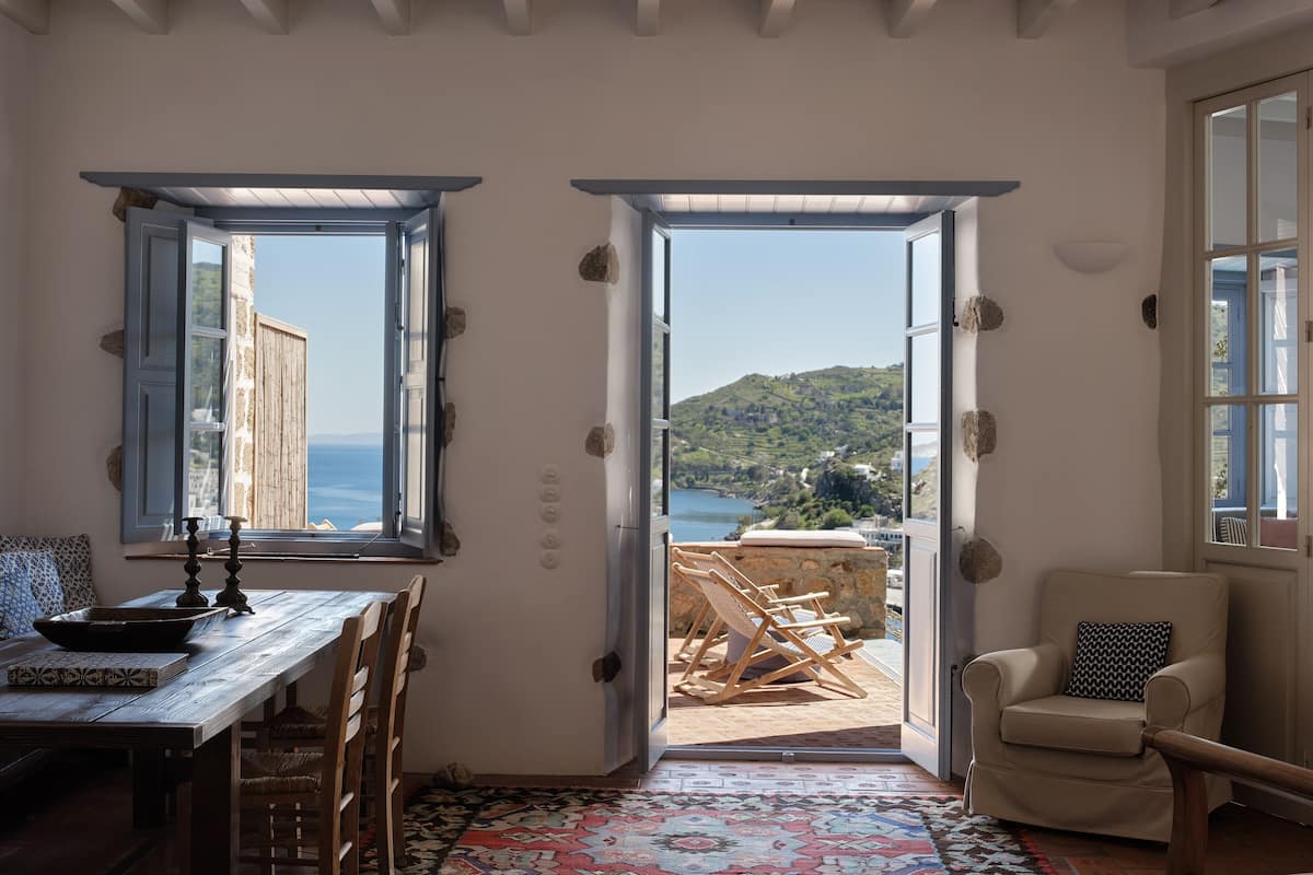 A room with a wooden table, chairs, a patterned rug, and a view of a hillside through open doors.