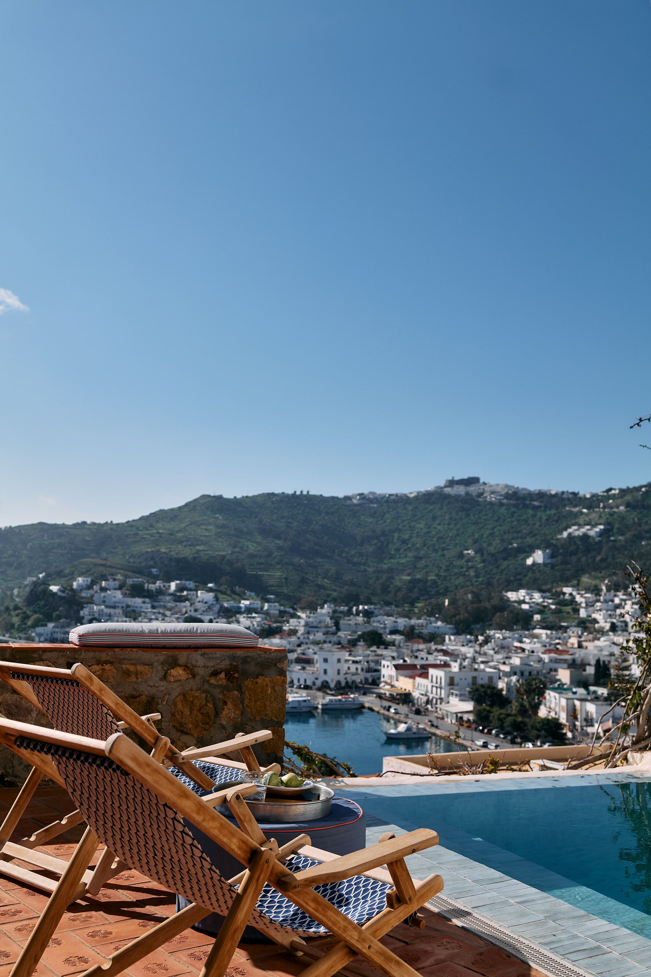 A poolside area with lounge chairs, a stone wall, and a view of a hillside town.