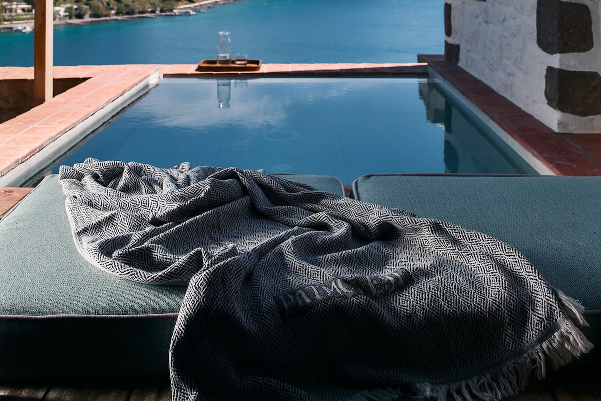 A pool with a view of a body of water and hills, a pergola, and a stone wall.