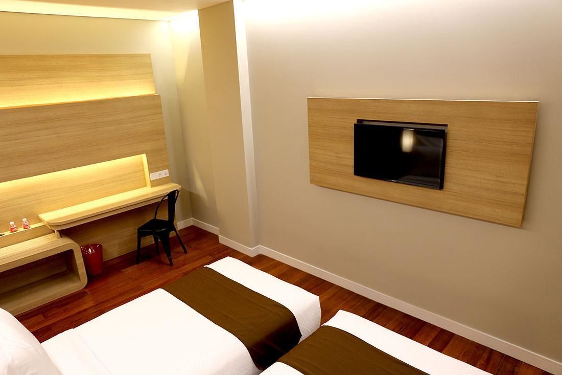 A hotel room with two beds, a desk with a chair, a TV mounted on the wall, and a wooden bench.