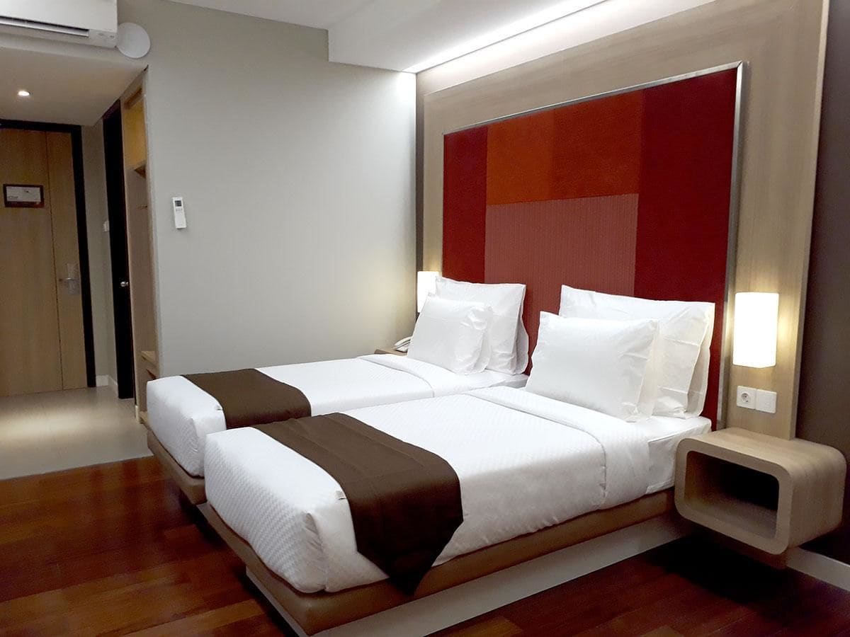 Een hotelkamer met twee bedden, een houten vloer, een rode accentmuur en een nachtkastje met een lamp.