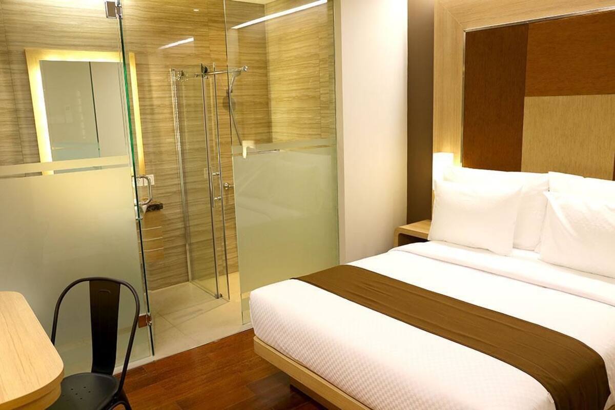 Ein modernes Hotelzimmer mit einem großen Bett, einer gläsernen Dusche, Holzboden und einem kleinen Tisch mit Stuhl.