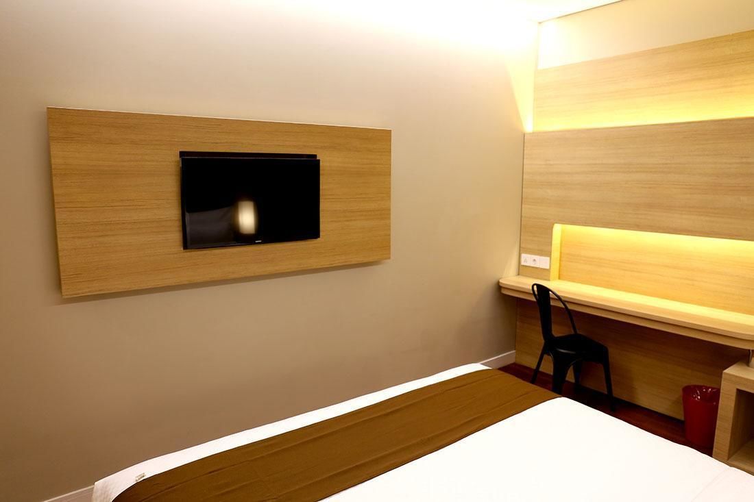 A hotel room with a bed, a TV mounted on a wooden panel, a desk with a chair, and a red trash bin.