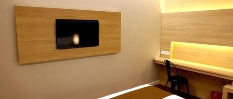 A hotel room with a bed, a TV mounted on a wooden panel, a desk with a chair, and a red trash bin.