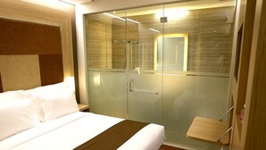 A modern hotel room with a bed, a glass-enclosed walk-in closet, and a small wooden shelf. - Grand Citihub Hotel at Panakkukang (Makassar)