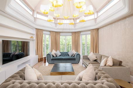 A spacious living room with a large sofa, a smaller sofa, and a coffee table. There is a television mounted on the wall, and the room has large windows with curtains.