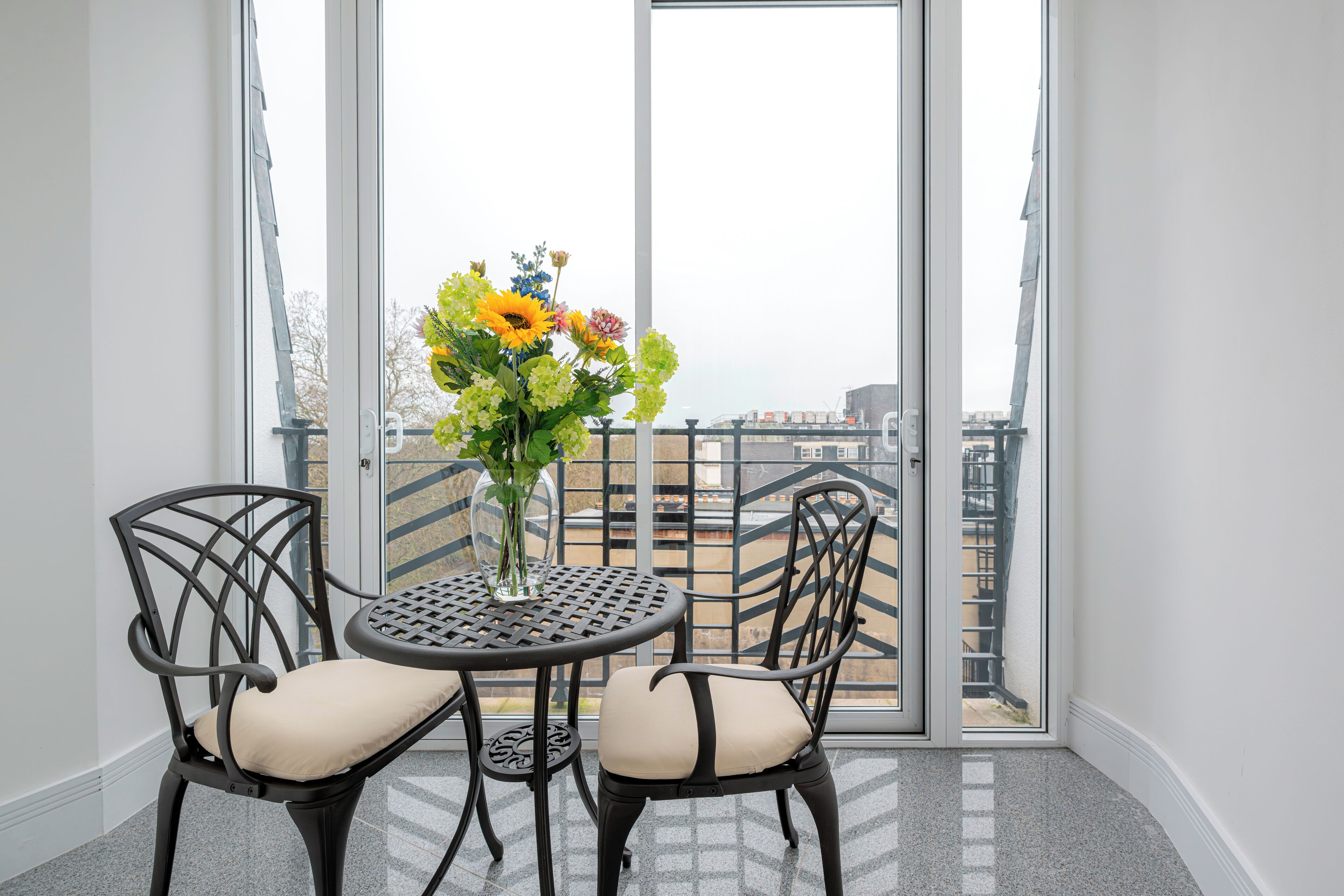 En balkon med et bord og stole, en vase med blomster og udsigt til bygninger.