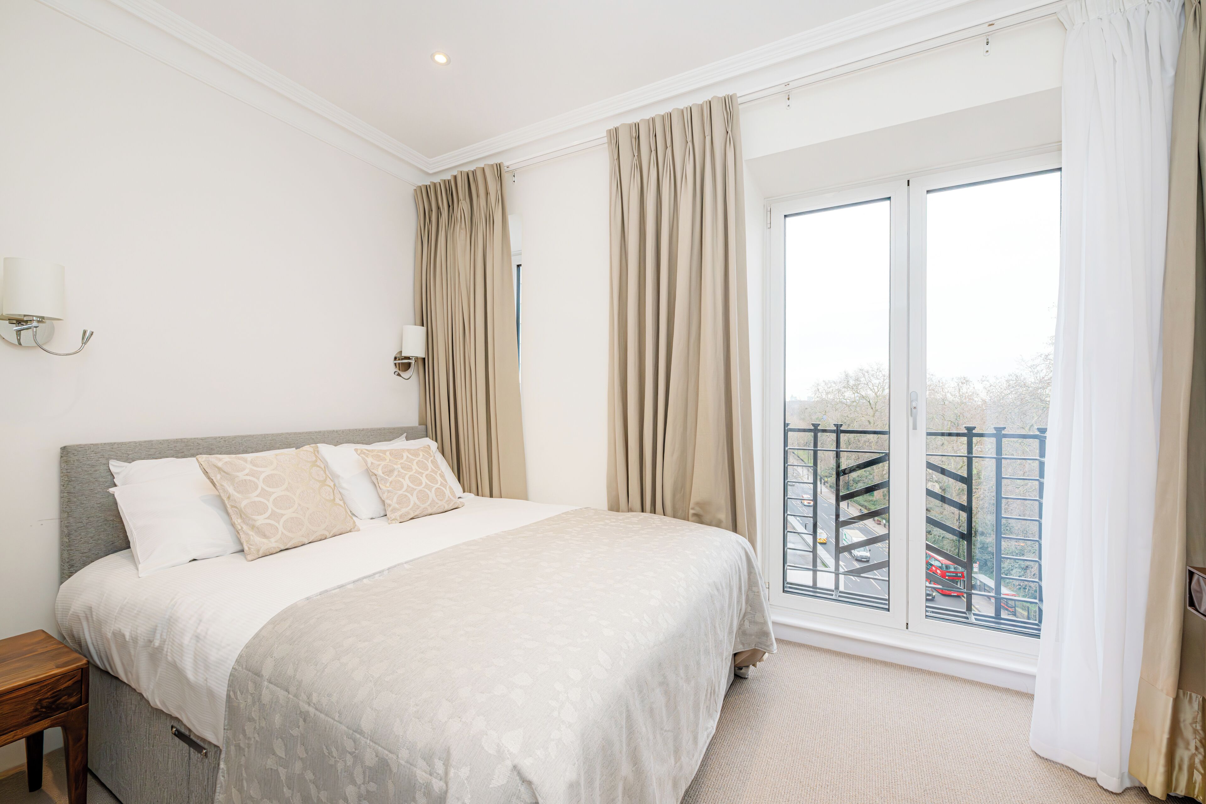A bedroom with a large bed, a nightstand, a window with curtains, and a view of a balcony.