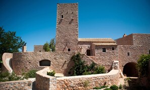 Un bâtiment historique en pierre, doté d’une tour imposante, de fenêtres en arc et d’une cour aménagée en terrasses.