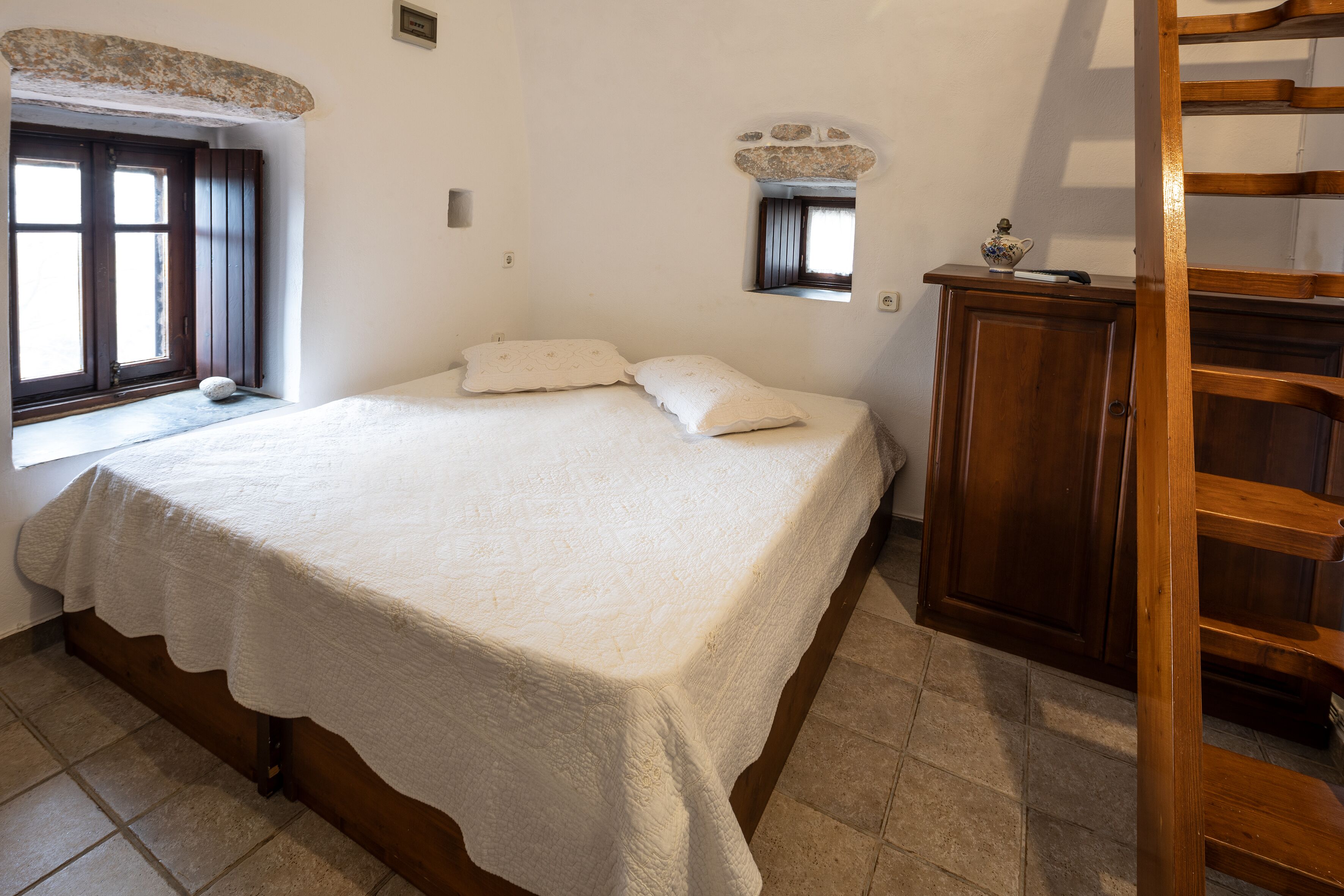 A bedroom with a large bed, a wooden wardrobe, a window, and a small mirror.