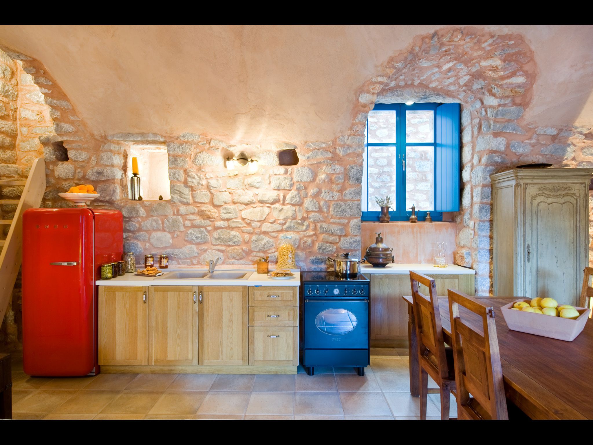 A rustic kitchen with stone walls, a red refrigerator, wooden cabinets, a blue dishwasher, and a dining area with a table and chairs.
