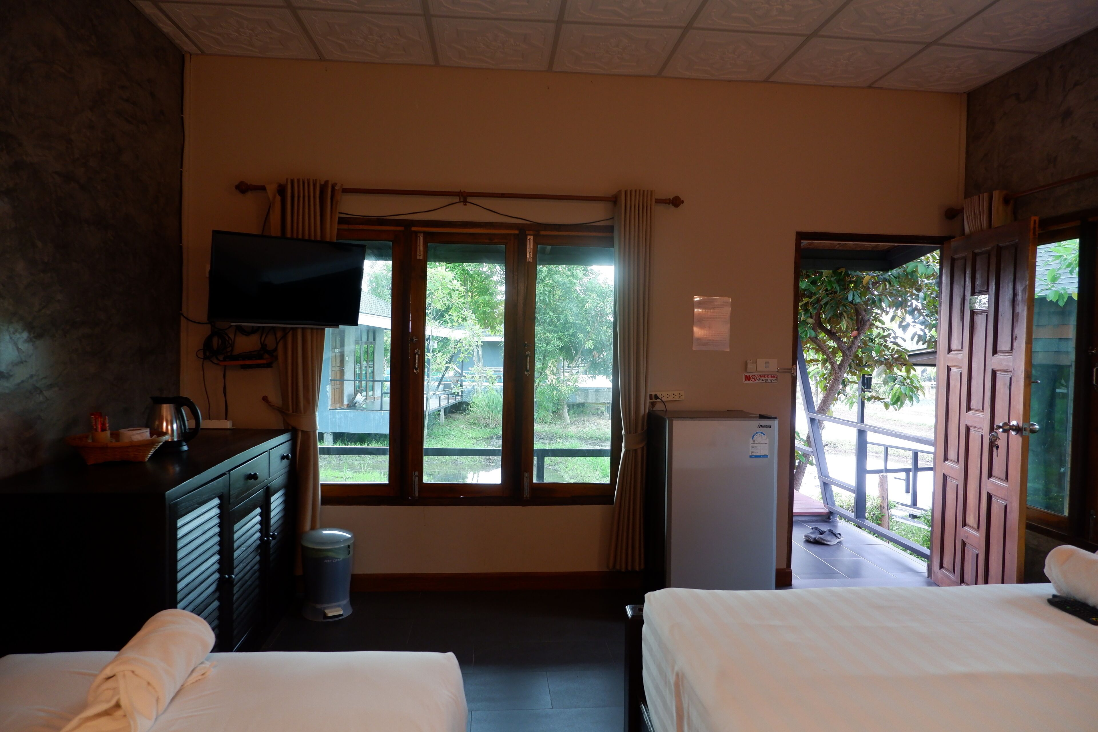 A hotel room with two beds, a TV, a desk with a kettle, a trash bin, and a view of the outdoors.