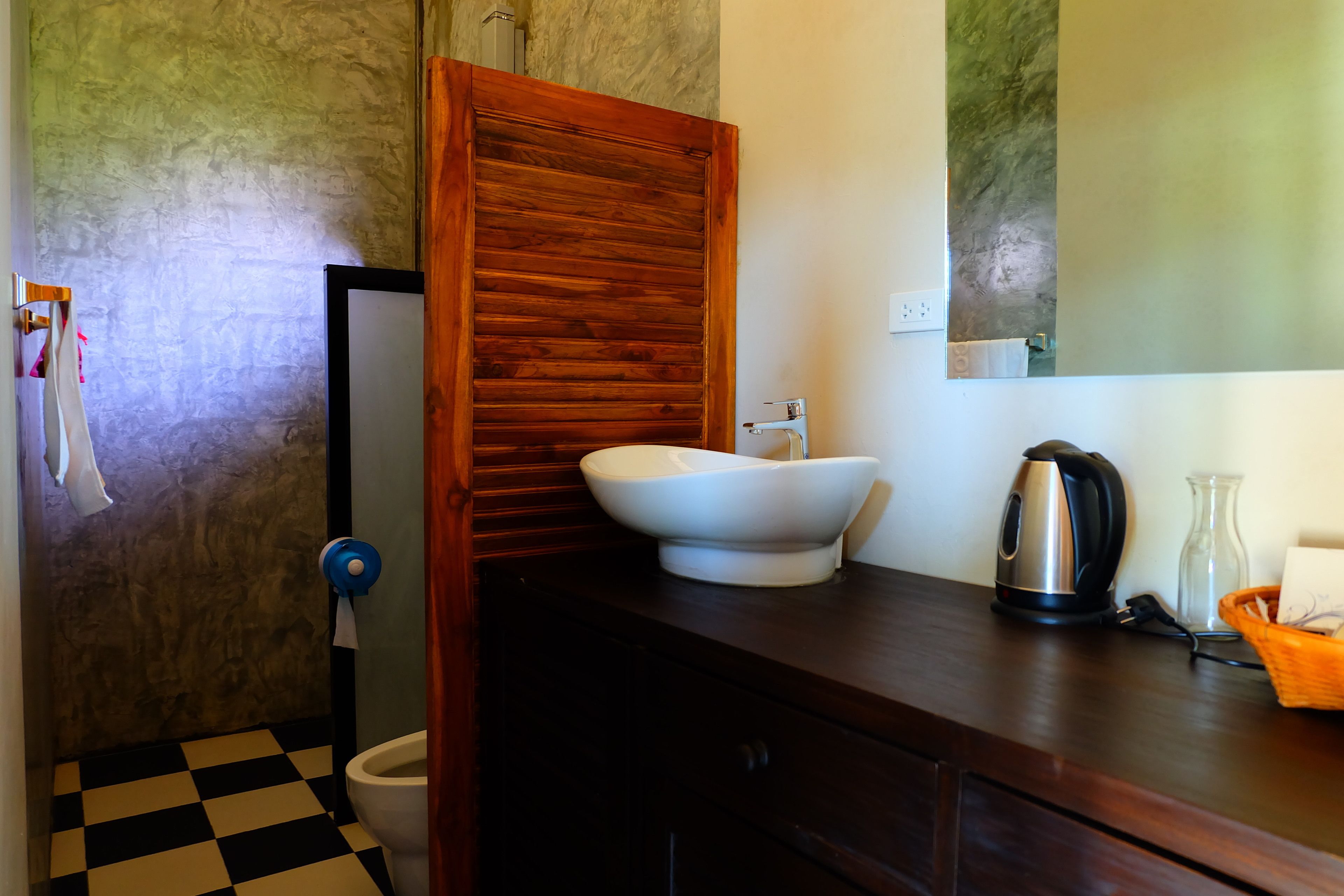 Une salle de bain avec un carrelage à motifs, une porte en bois, un lavabo blanc, une bouilloire et un pichet en verre.