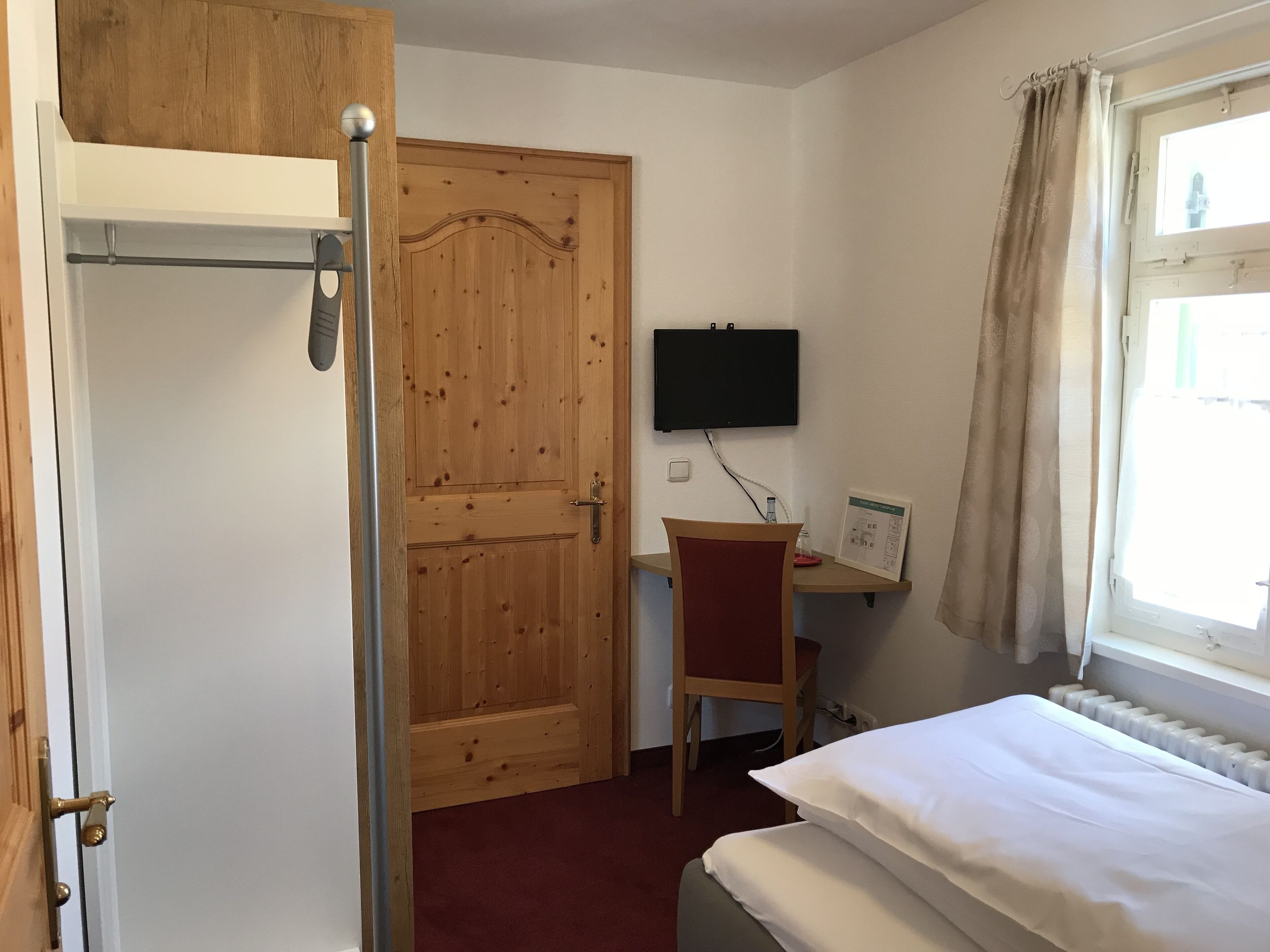 Ein Zimmer mit Holztür, einem Bett, einem Schreibtisch mit Stuhl, einem an der Wand befestigten Fernseher und einem Fenster mit Vorhängen.