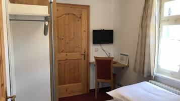 Ein Zimmer mit Holztür, einem Bett, einem Schreibtisch mit Stuhl, einem an der Wand befestigten Fernseher und einem Fenster mit Vorhängen.
