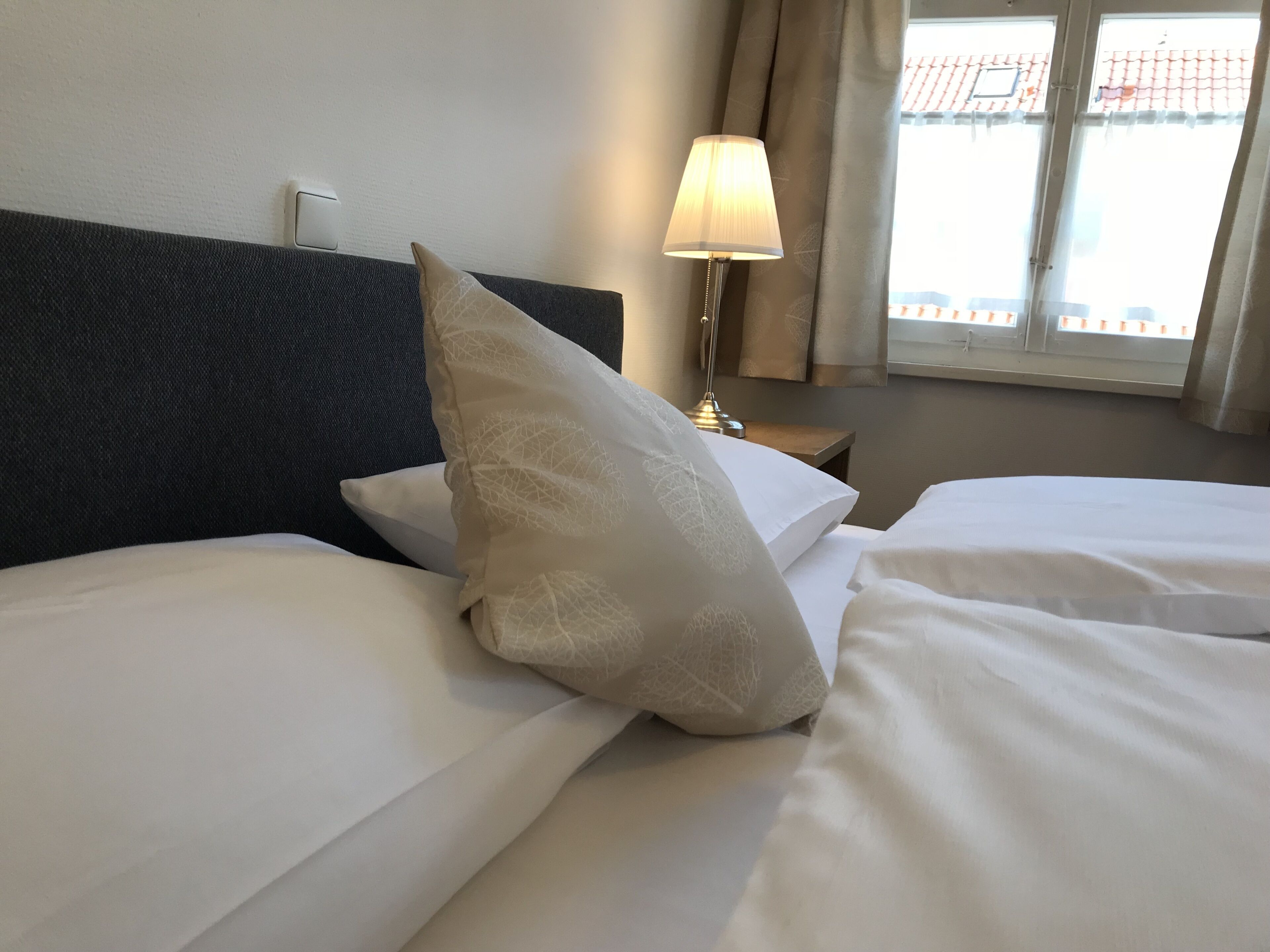 Habitación de hotel con dos camas, cabecero oscuro, lámpara de noche y ventana con cortinas.