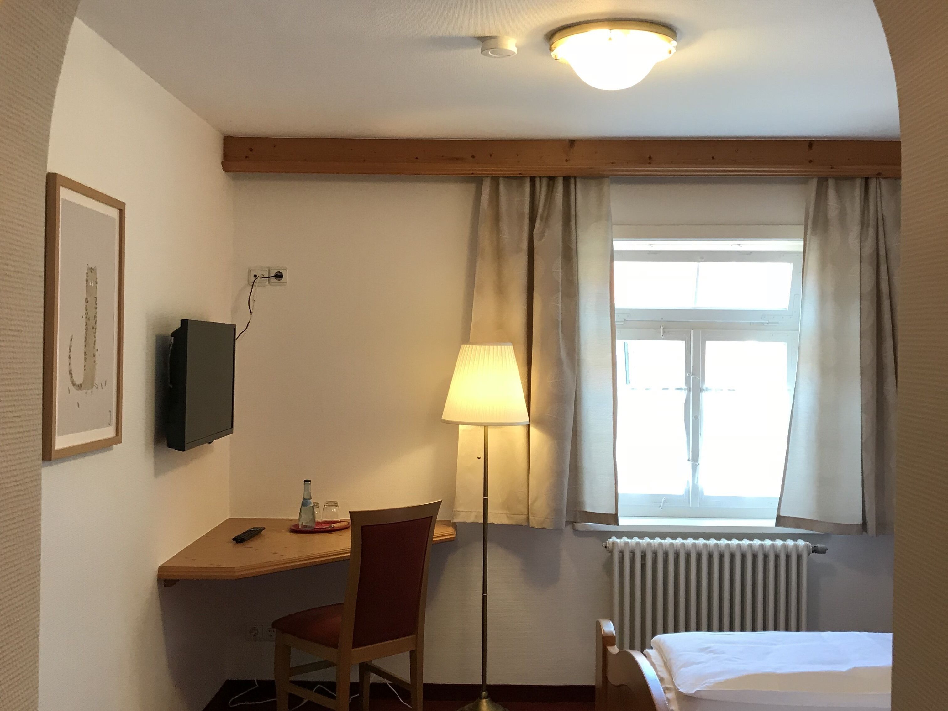 Una habitación de hotel con escritorio, silla, lámpara, cama, ventana con cortinas y radiador.