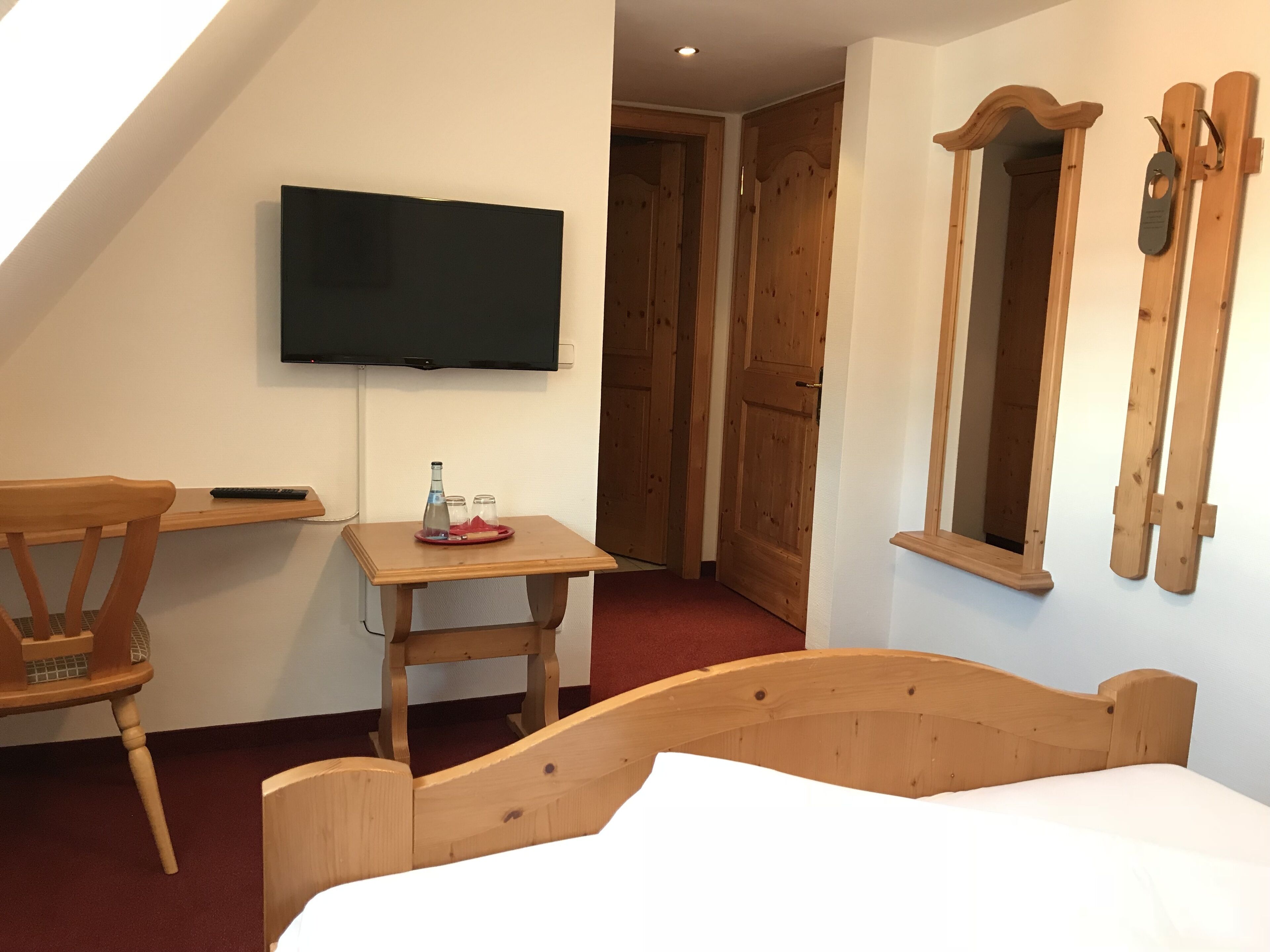 Ein Zimmer mit einem Bett, einem an der Wand befestigten Fernseher, einem kleinen Holztisch mit Flasche und Gläsern sowie einem Spiegel.
