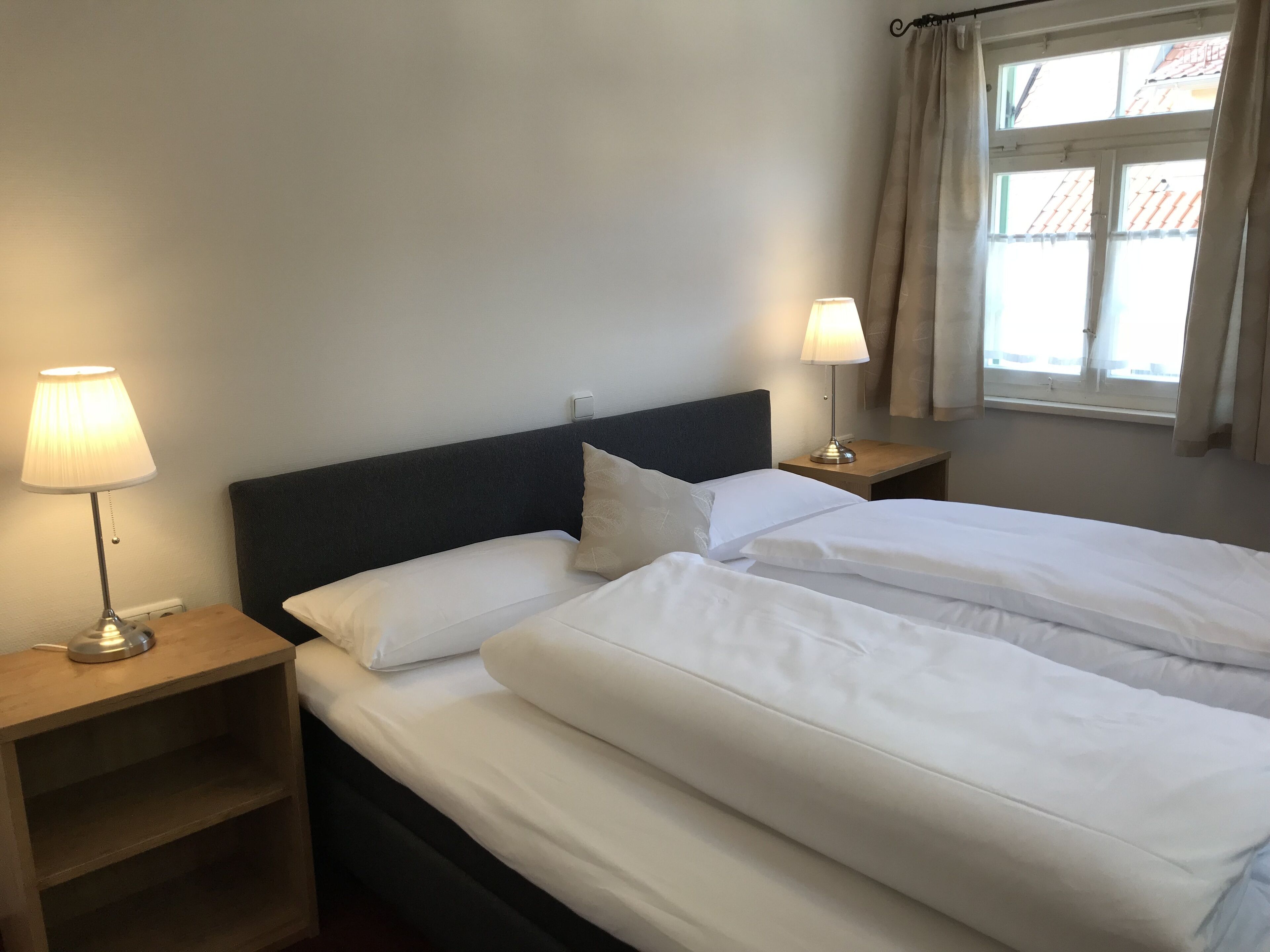 A double bed with white linens, two bedside tables with lamps, a dark headboard, and a window with curtains.