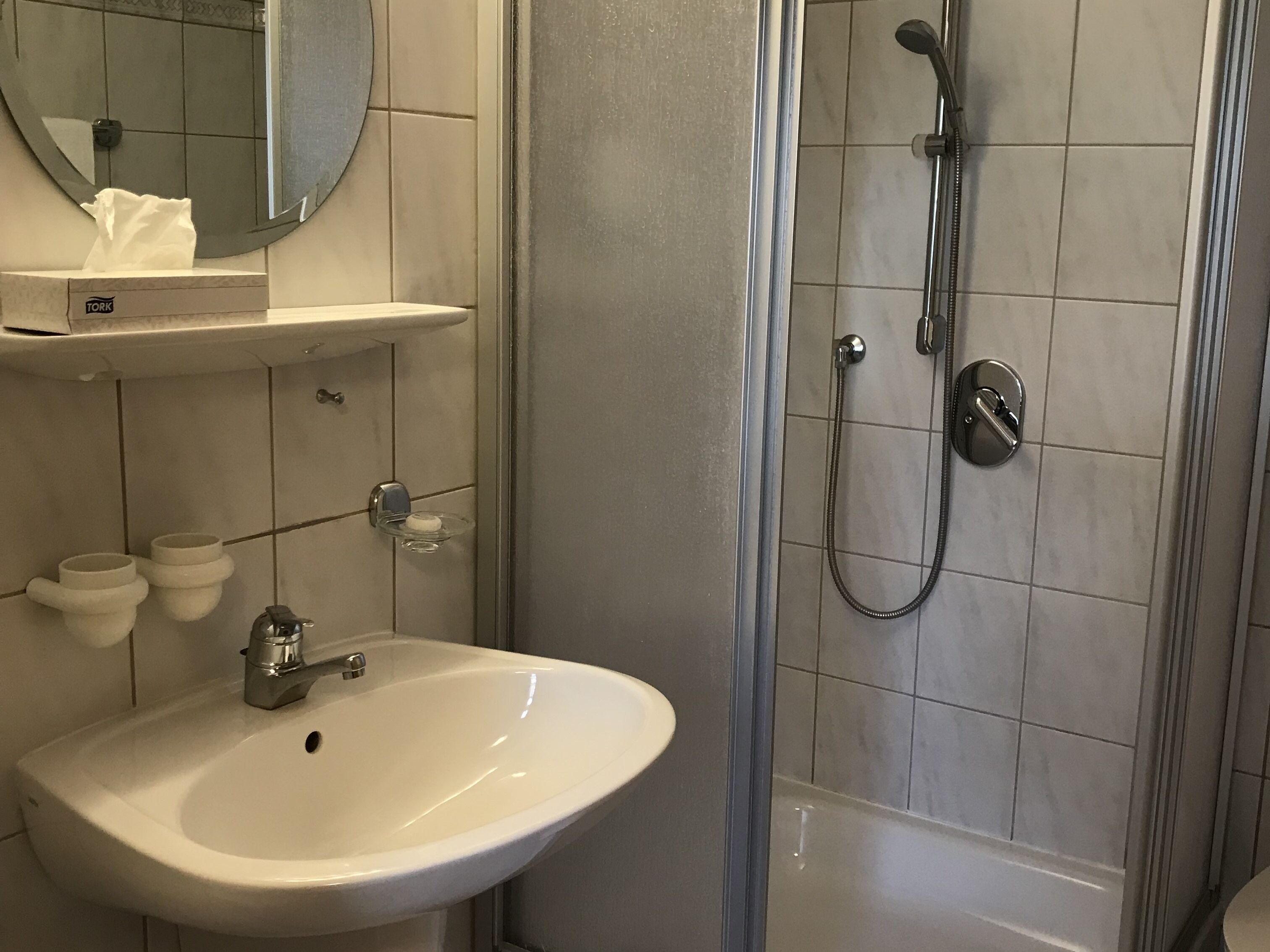 A bathroom with a white sink, a mirror, a towel dispenser, and a shower with a handheld showerhead.