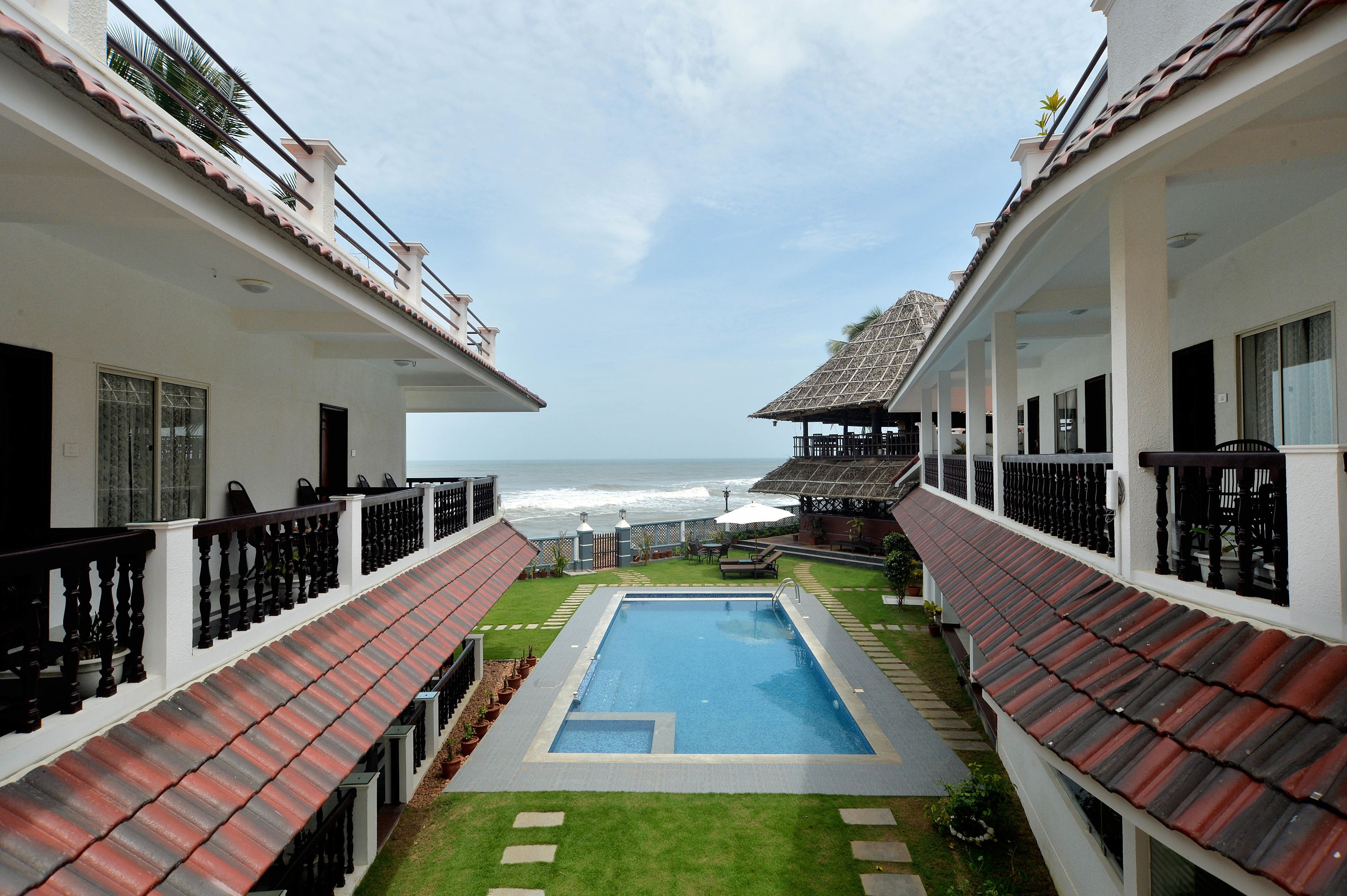 A pool area with a view of the ocean, surrounded by buildings with balconies and a thatched roof structure.