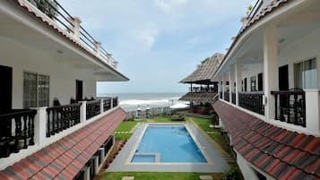 A pool area with a view of the ocean, surrounded by buildings with balconies and a thatched roof structure.