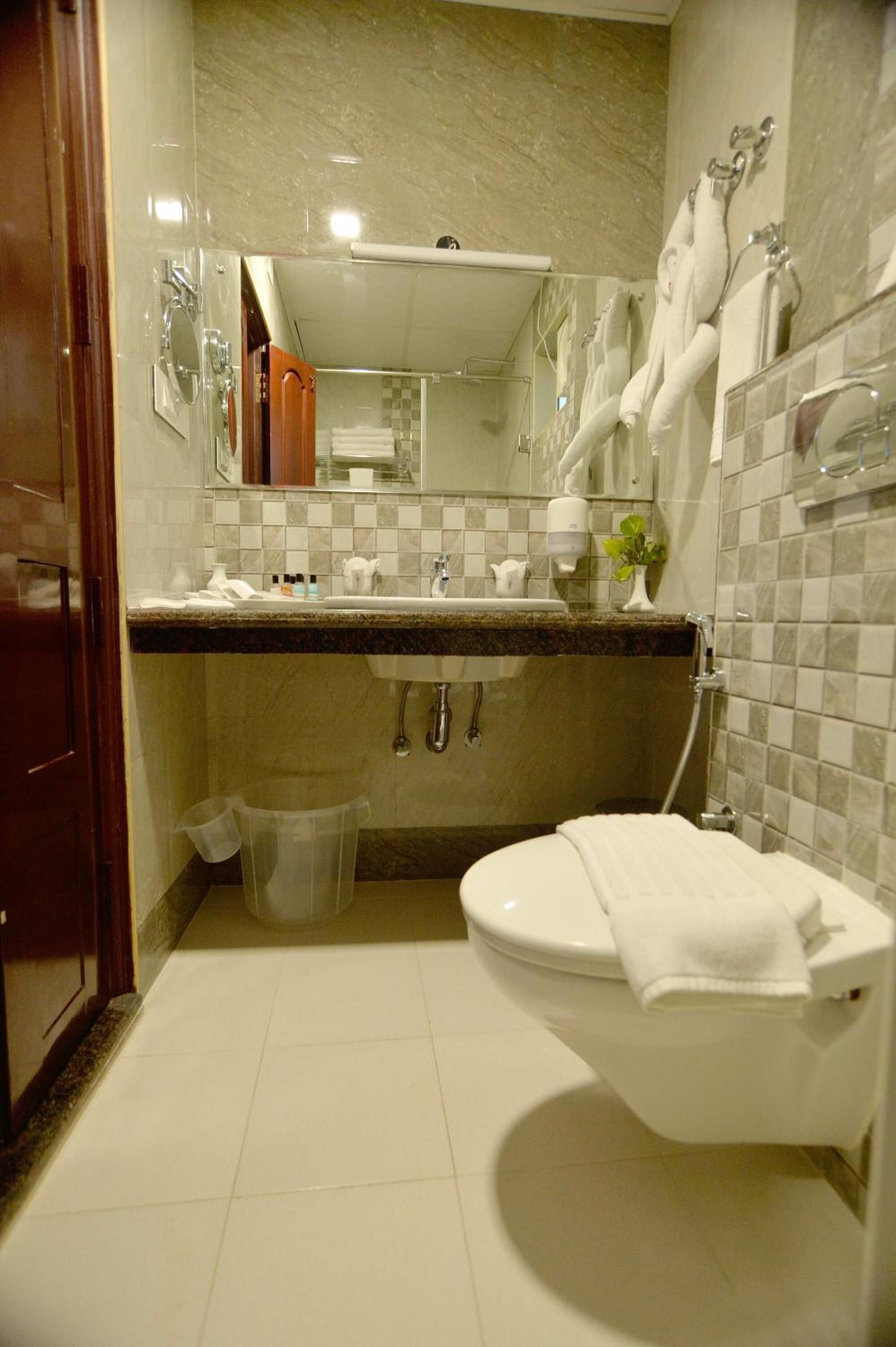 A bathroom with a toilet, sink, and mirror. There are towels hanging on the wall and a trash can.
