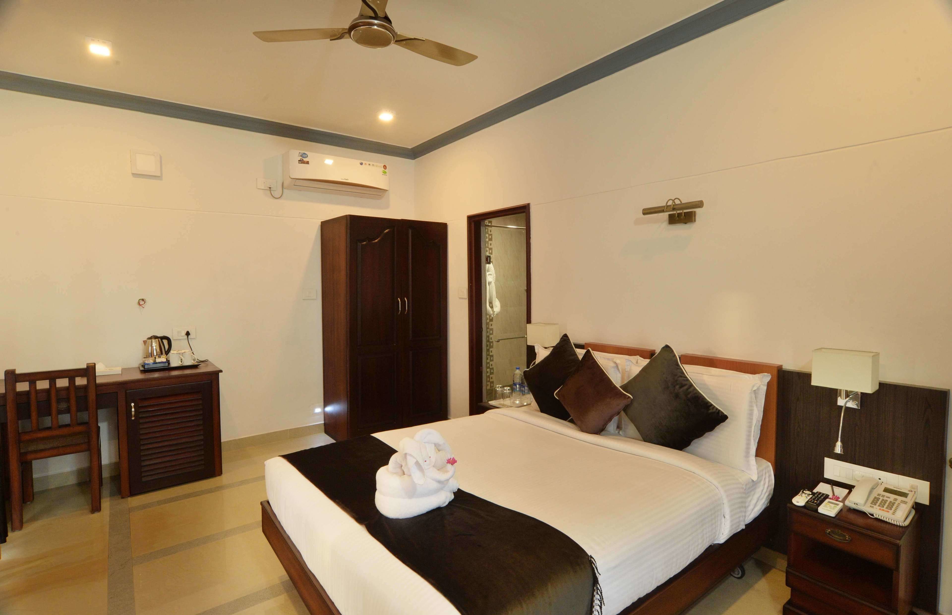 A hotel room with a bed, bedside table, desk, chair, wardrobe, and ceiling fan.