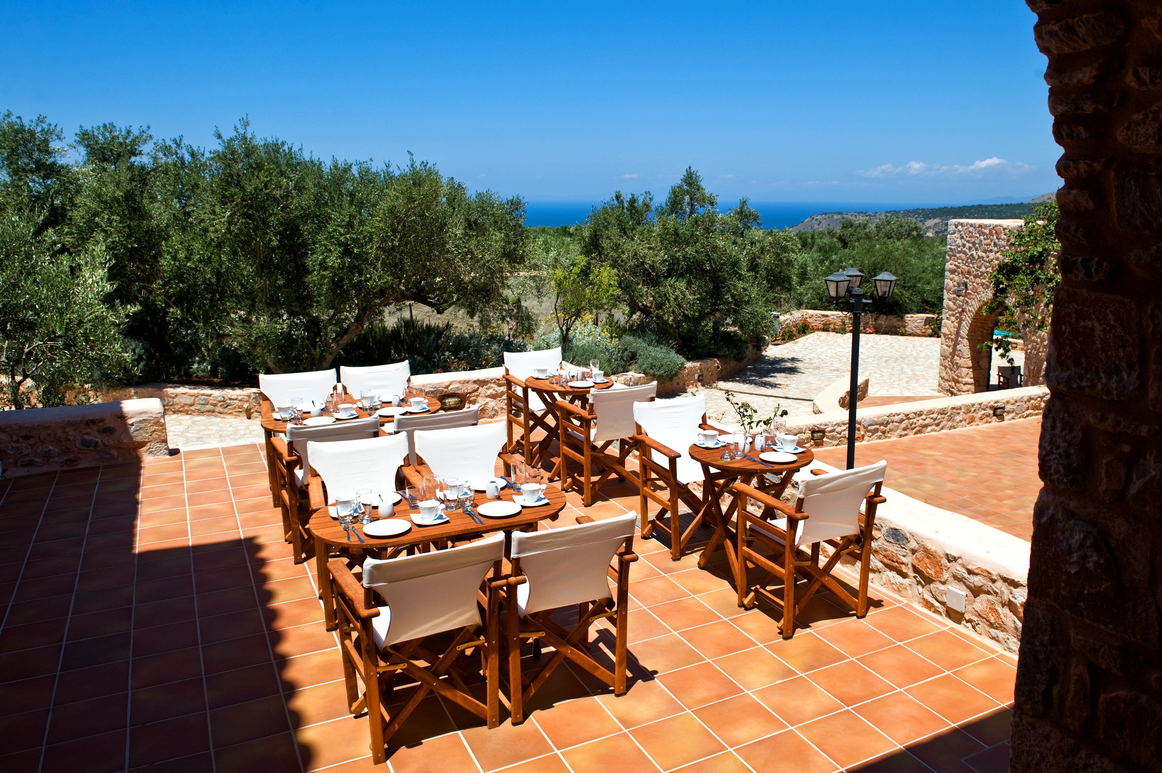 An outdoor dining area with multiple tables and chairs, set against a backdrop of trees and a view of the sea.