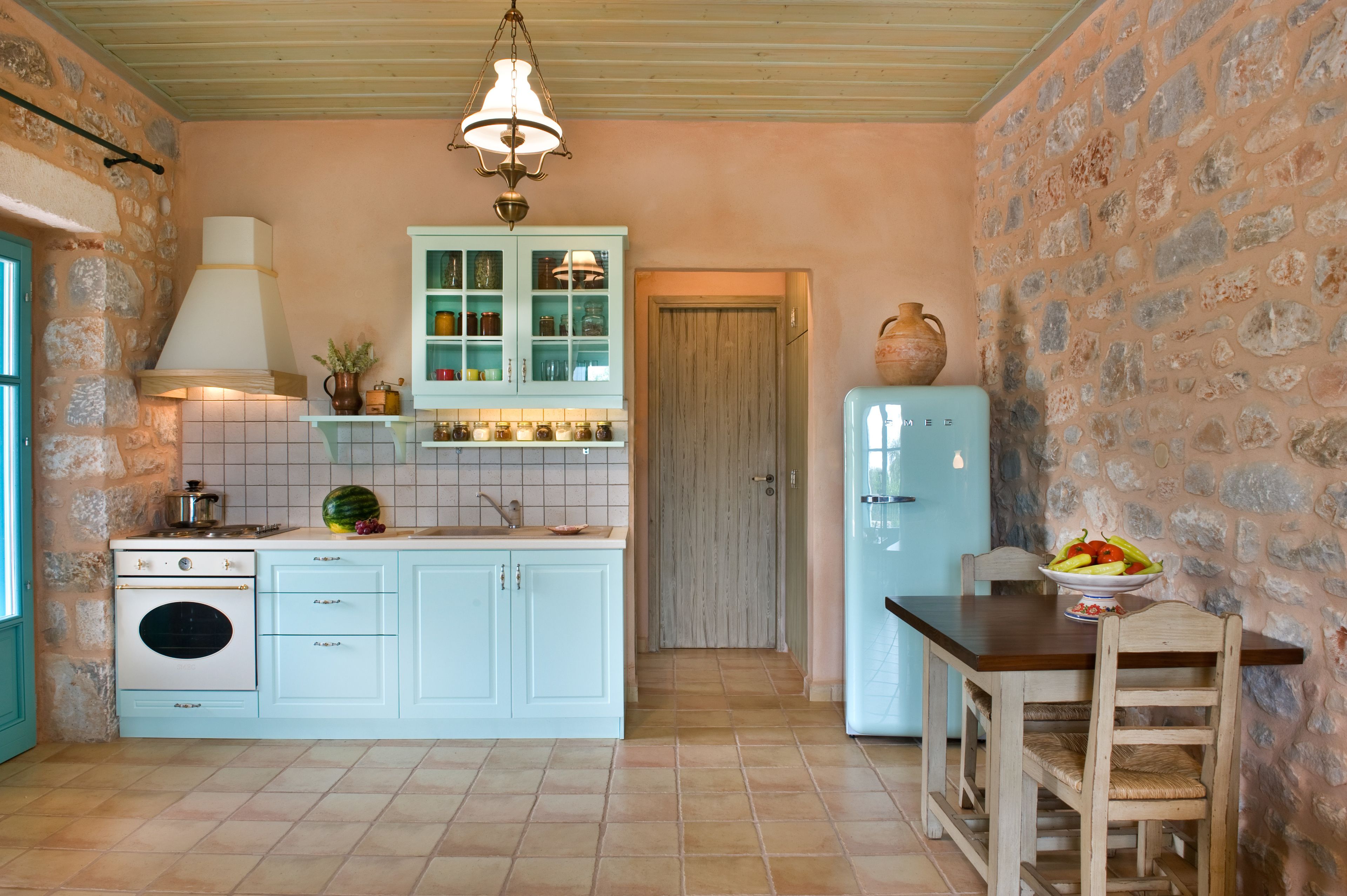 Una cocina con pared de piedra, estufa blanca, armario azul claro y mesa de madera con sillas.