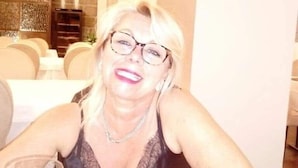 Une personne est assise à une table, un verre de vin à la main, dans une pièce dont le mur est en pierre et qui est ornée d’un lustre.