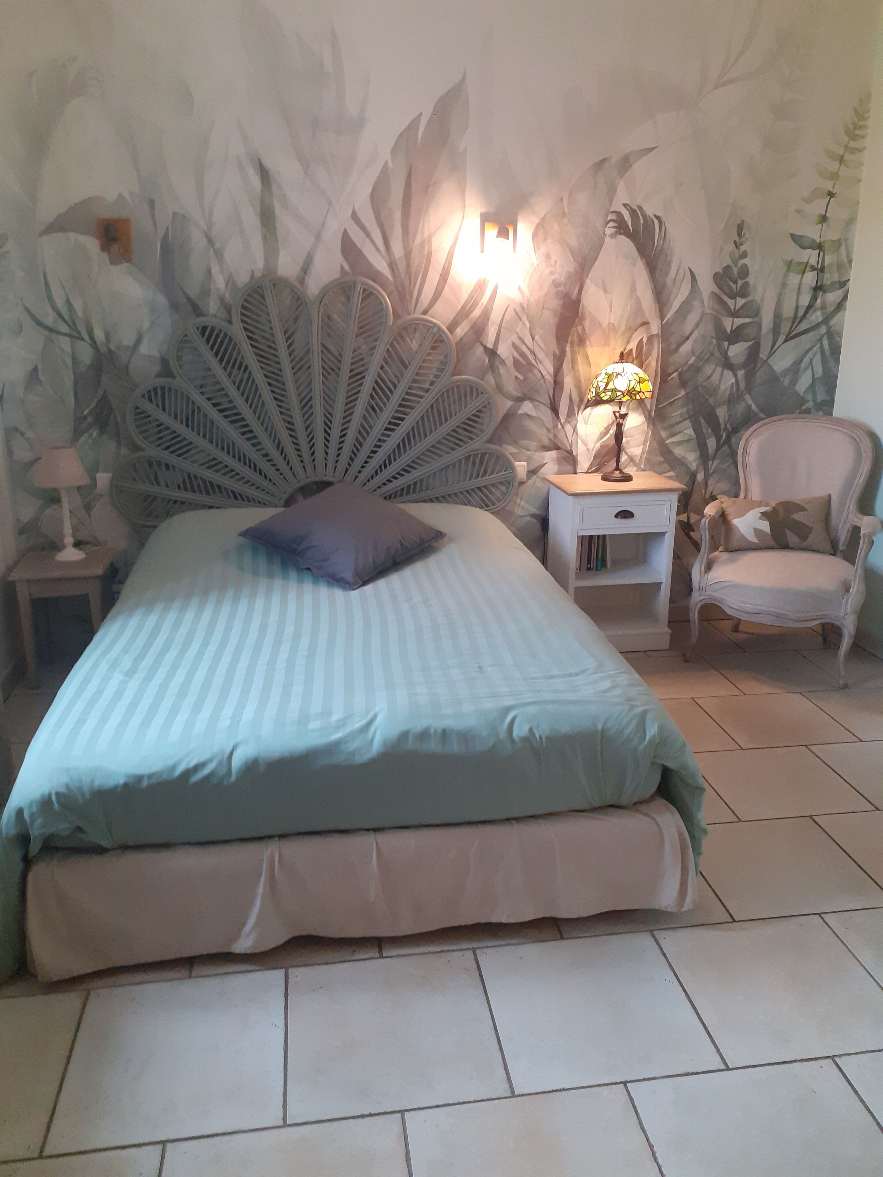 A bedroom with a bed, a nightstand, a lamp, a chair, and a wall with a leaf pattern.