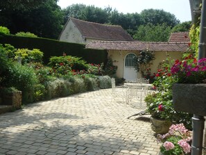 Un chemin de jardin pavé qui mène à un bâtiment doté d’un toit en tuiles, entouré d’une végétation luxuriante et de plantes en fleurs.
