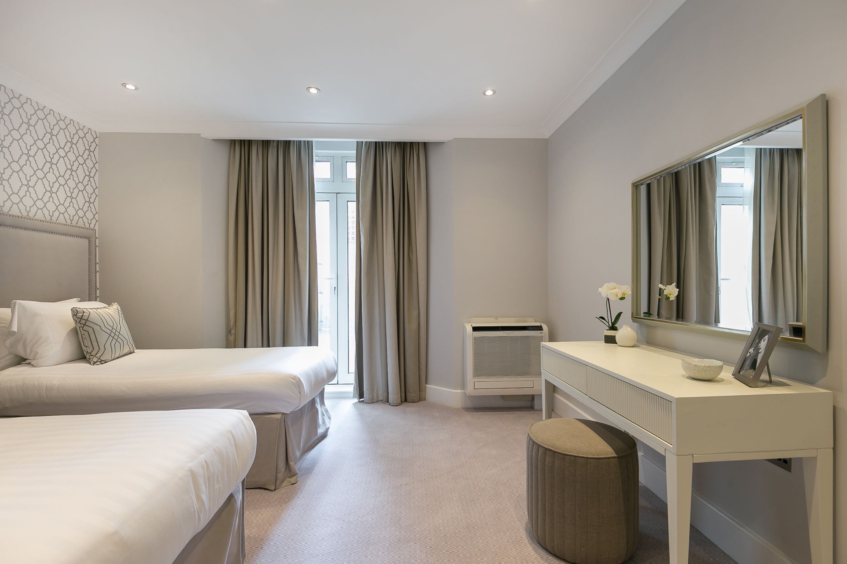 A hotel room with two beds, a vanity with a mirror, and a window with curtains.