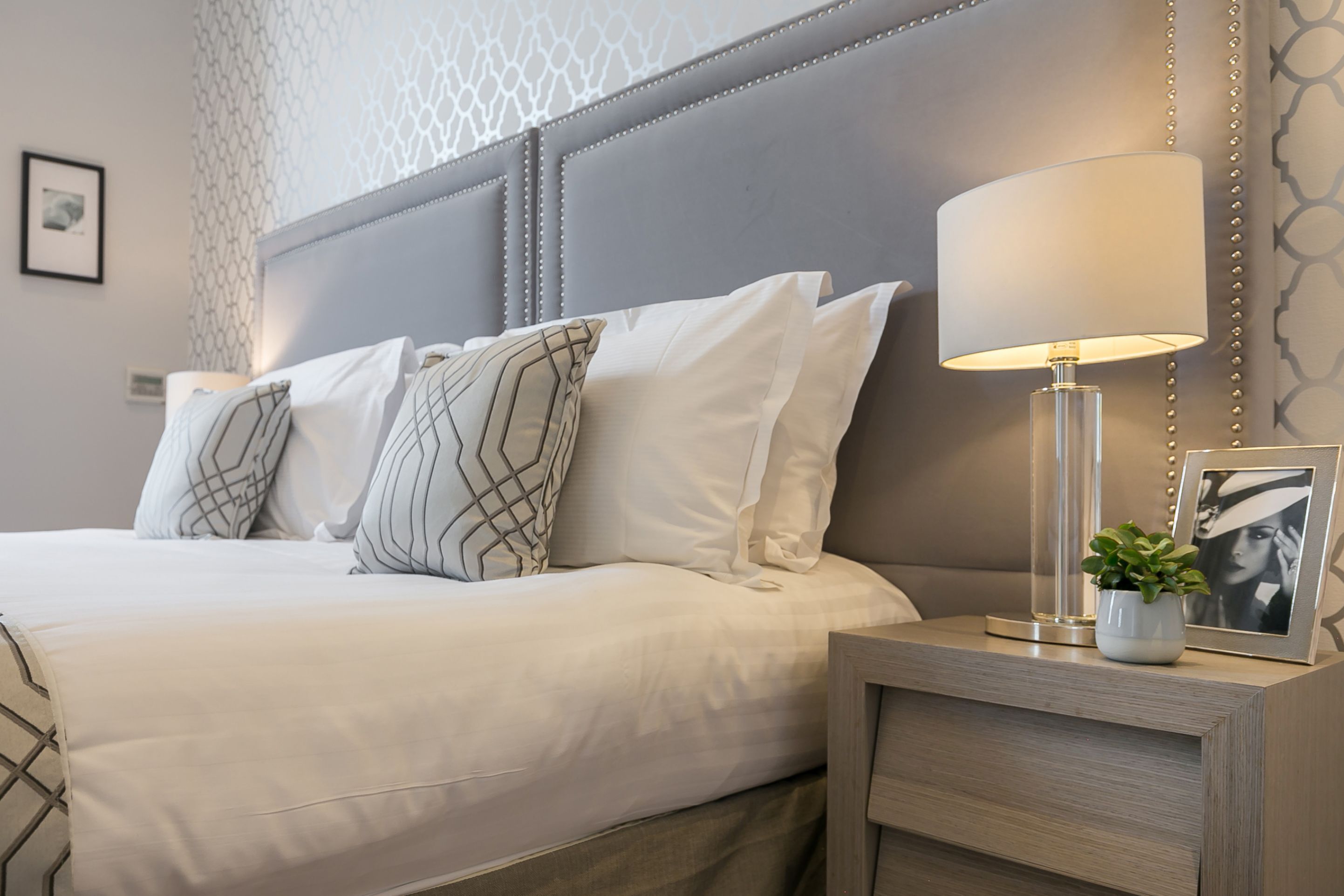A neatly made bed with white linens and patterned pillows, a bedside table with a lamp and a framed picture, and a decorative wall panel.