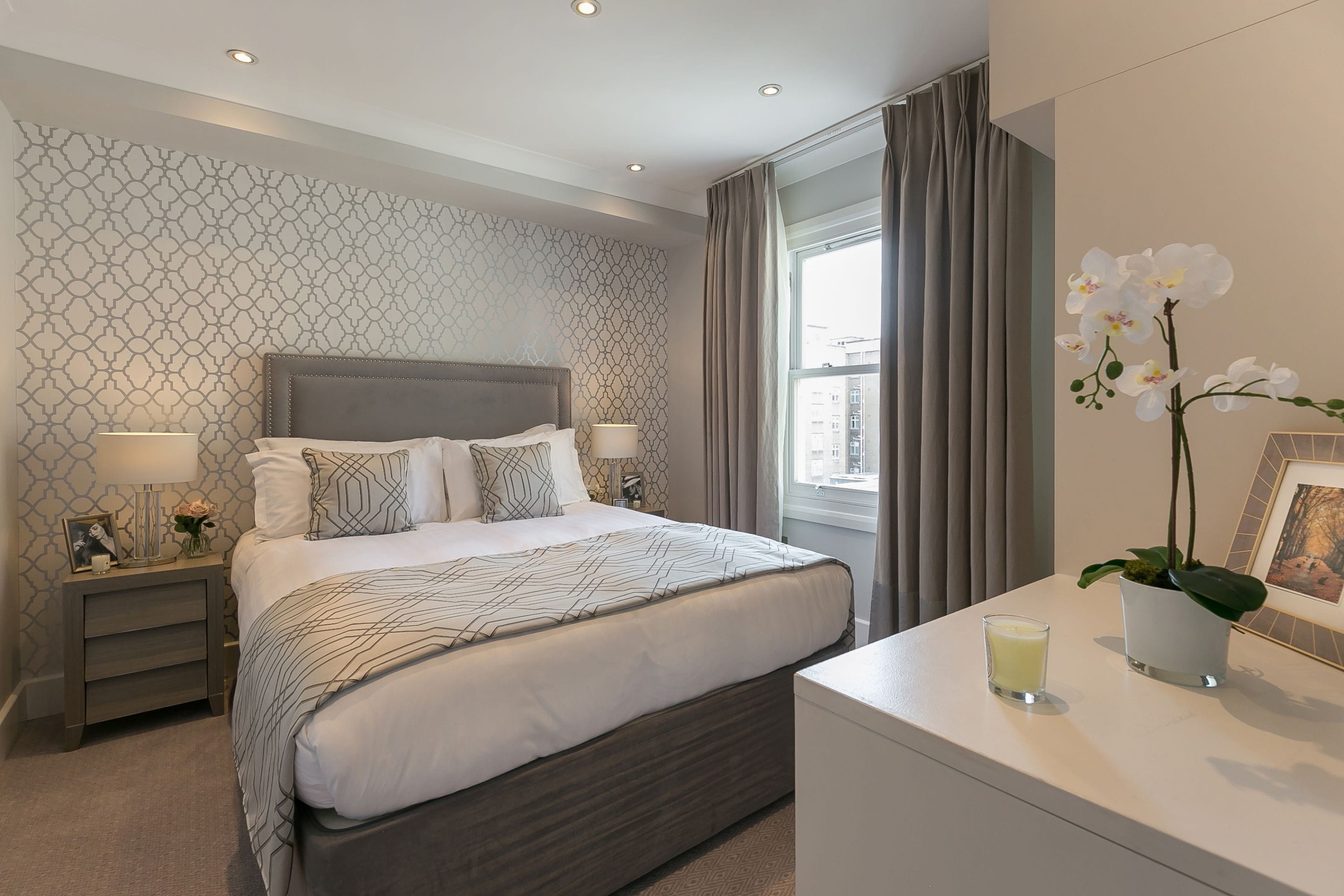 A neatly arranged bedroom with a large bed, bedside tables, a bedside lamp, a small table with a candle, and a potted orchid.